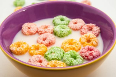 Süt içinde renkli kahvaltı gevrekleri olan, yenmeye hazır bir kase ve masada biraz mısır gevreği. Resim yatay formatta.