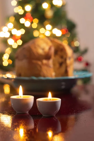 A defocused panettone with two lit candles and the Christmas tree lights in the background. Typical Christmas food.