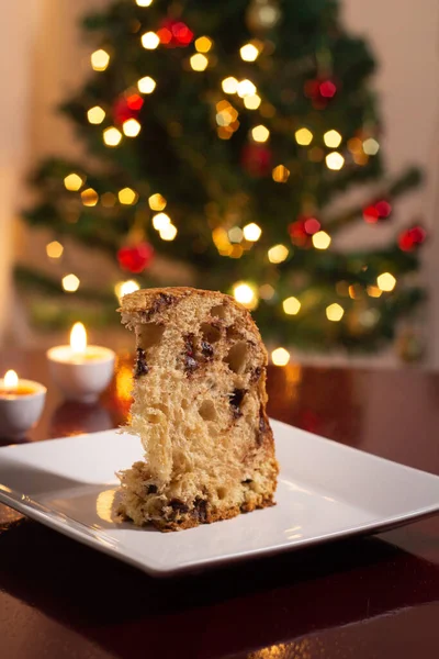 A slice of panettone inside the plate, two lit candles and the lights of the Christmas tree in the background. Typical Christmas food.