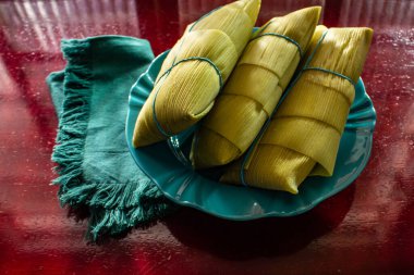 Parlak bir yüzey üzerinde, bir tabağın içinde, mısır kabuklarıyla kaplı 3 pamonha..