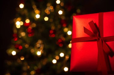 Kırmızı bir hediye kutusu, kurdeleli, arkasında Noel ağacı olan..