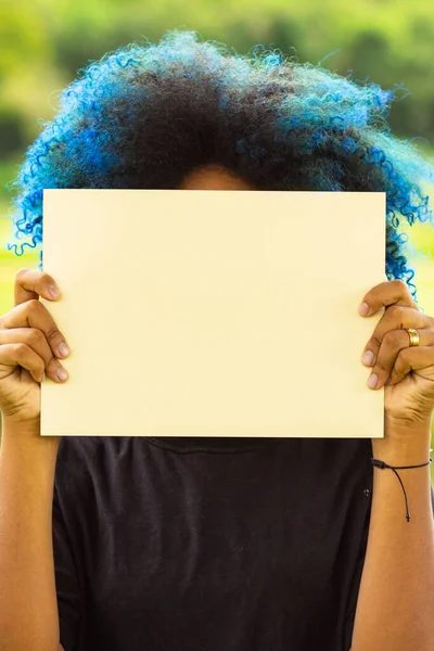 A young woman, with hair dyed blue, her face hidden behind a blank poster, with a landscape in the background.