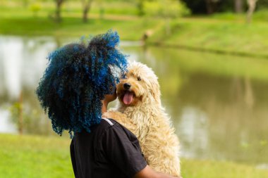 Uma jovem de cabelos tingidos de azul, abraando carinhosamente seu cachorro no colo, com um parque arborizado, desfocado ao fundo.