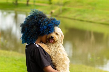 Uma jovem de cabelos tingidos de azul, abraando carinhosamente seu cachorro no colo, com um parque arborizado, desfocado ao fundo.