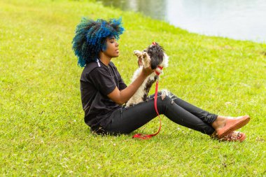 Genç siyah bir kadın, kıvırcık saçları maviye boyanmış, parkta çimenlerde oturuyor, kucağındaki köpeğiyle oynuyor..