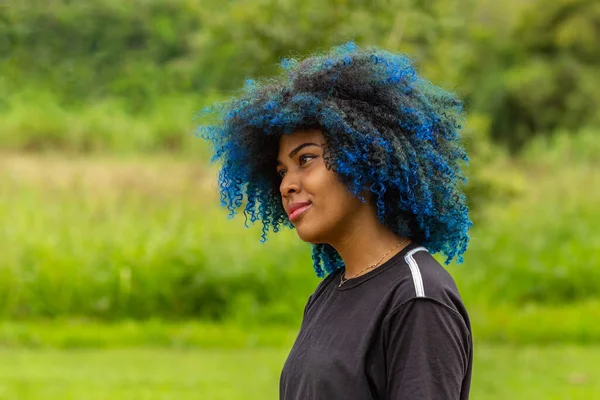 Photo of a young black woman with afro hair, dyed blue, with a slight smile on her face, in profile with blurred park vegetation in the background.