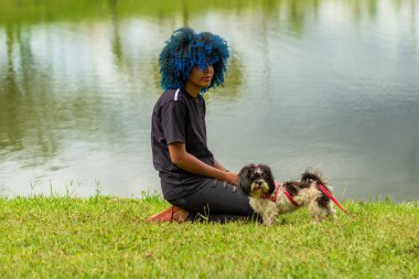 Genç siyahi bir kadın, köpeğiyle gölün kenarında çimlerde oturuyor..