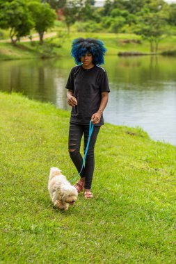 Genç siyah bir kadın, saçları maviye boyanmış, çimenlikte bir köpek gezdiriyor, parkta bir göletin yanında..