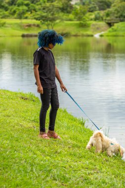 Genç siyah bir kadın, saçları maviye boyanmış, çimenlikte bir köpek gezdiriyor, parkta bir göletin yanında..