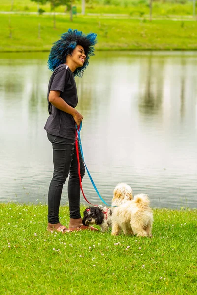 A young black woman, with afro hair dyed blue, walking her dogs on the lawn beside a pond in the park.