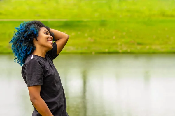 A young black woman, happy, in a very green natural park, straightening her dyed blue hair.
