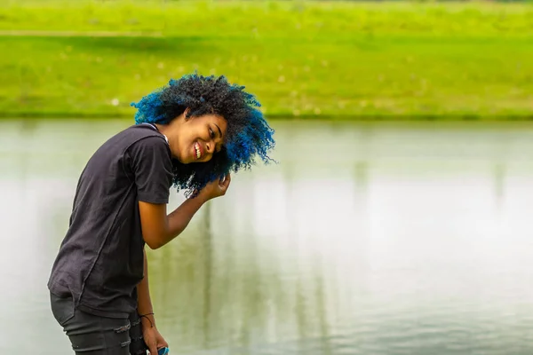 A young black woman, happy, in a very green natural park, straightening her dyed blue hair.