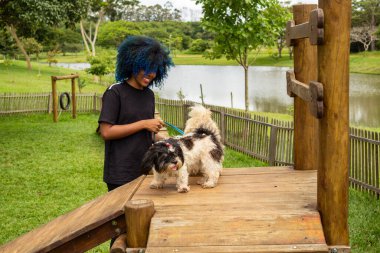 Mavi saçlı genç bir kadın, bir sürü ağaç ve gölü olan bir parktaki ahşap bir binada köpekleriyle oynuyor..