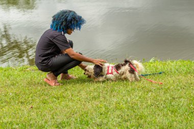 Siyahi bir kadın, boyalı mavi saçlı, çimlere çömelmiş, köpeğiyle arka planda bir gölle birlikte ağaçlık bir parkta oynuyor..