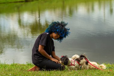 Siyahi bir kadın, boyalı mavi saçlı, çimlere çömelmiş, köpeğiyle arka planda bir gölle birlikte ağaçlık bir parkta oynuyor..