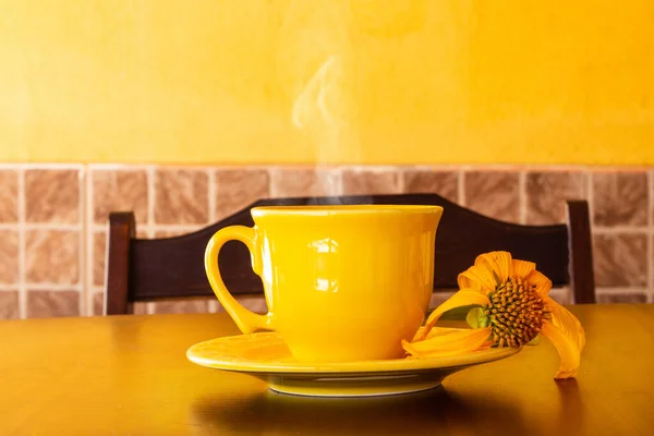 一杯黄色的咖啡 有茶托和一朵枯萎的向日葵 放在一张木制桌子上 后座上有一把椅子 图库图片