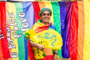 Sarı yelpazeli ve arka planında gökkuşağı bayrakları olan eşcinsel bir adam. Fotoğraf: Goiania 'daki LGBTQIA + Gurur Yürüyüşü.