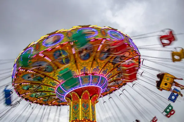 A giant spinning ride, spinning, with chairs attached by chains flying high, with few people, in an amusement park, on a cloudy day. Photo taken from bottom to top at low camera speed.