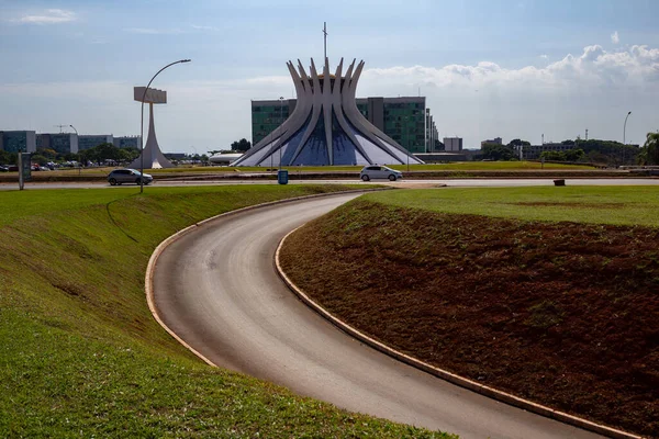 Güneşli bir sabahta Brasilia Metropolitan Katedrali ve açık gökyüzü olan eğimli, asfalt bir bulvar. 