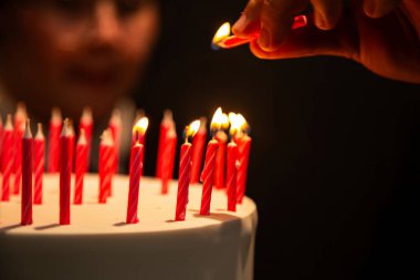 Arka planda bulanık bir yüz olan doğum günü pastasında mum yakan bir elin ayrıntıları.