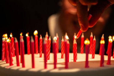 Arka planda bulanık bir yüz olan doğum günü pastasında mum yakan bir elin ayrıntıları.