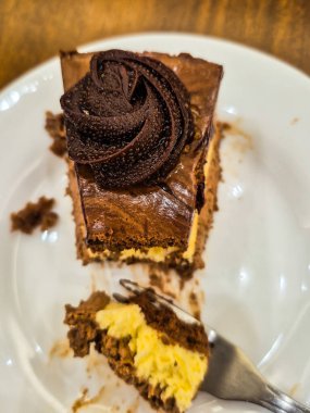 Beyaz bir porselen tabağın içinde kremalı bir parça çikolatalı pasta. Pastanın bir kısmı çatallı, yemeye hazır..