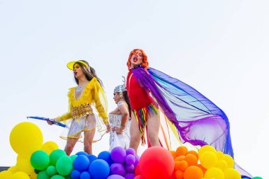 LGBTQIA + Gurur Yürüyüşü sırasında, renkli balonlarla süslenmiş, açık renkli ve dalgalı elbiseler giyen, elektrikli üçlü kamyonda dans eden üç kişi..
