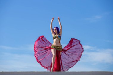 Kırmızı kıyafetli bir dansçı, açık etekli, ışıktan yapılmış, dalgalı kumaşlı, arkasında mavi gökyüzü olan..