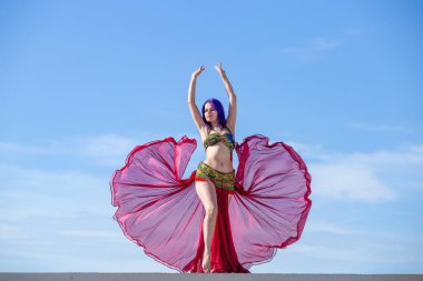 Kırmızı kıyafetli bir dansçı, açık etekli, ışıktan yapılmış, dalgalı kumaşlı, arkasında mavi gökyüzü olan..