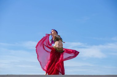 Kırmızı elbiseli, parlak etekli, akıcı kumaşlı, arka planda mavi gökyüzü olan bir dansçı..