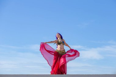 Kırmızı elbiseli, parlak etekli, akıcı kumaşlı, arka planda mavi gökyüzü olan bir dansçı..