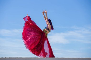 Kırmızı elbiseli bir dansçı, bacağı havada hareket ediyor, çok dolgun bir etek giyiyor ve arka planda mavi gökyüzü var..
