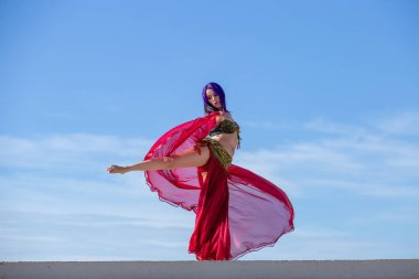 Arka planda mavi gökyüzü ile çelişen kırmızı kıyafetli bir dansçı. Akan eteğiyle zarif bir dönüş hareketi yapıyor..