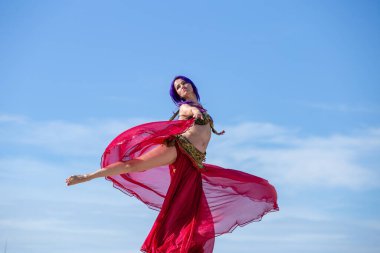 Arka planda mavi gökyüzü ile çelişen kırmızı kıyafetli bir dansçı. Akan eteğiyle zarif bir dönüş hareketi yapıyor..