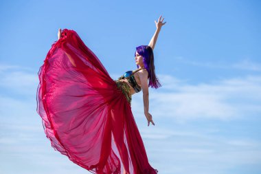 Kırmızı elbiseli bir dansçı, bacağı havada hareket ediyor, çok dolgun bir etek giyiyor ve arka planda mavi gökyüzü var..