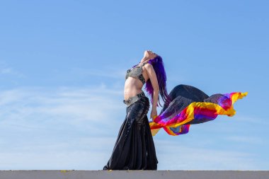 Dans eden bir kadın, göbek dansı, açık renk, dalgalı, arka planda mavi gökyüzü olan bir duvak..