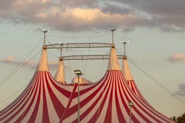 Sirk binasının tepesinde, kırmızı ve beyaz çizgili çadırlar ve ışık dizileri alacakaranlıkta, arka planda bulutlu bir gökyüzü ile aydınlandı..