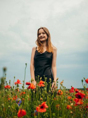 Bulutlu bir yaz gününde siyah bir gece elbisesi içinde gelincik tarlasına poz veren güzel bir genç kız. Açık havada bir kadın modelin portresi. Yağmurlu bir hava. Gri bulutlar. Dikey çekim.