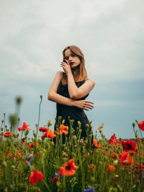 Bulutlu bir yaz gününde siyah bir gece elbisesi içinde gelincik tarlasına poz veren güzel bir genç kız. Açık havada bir kadın modelin portresi. Yağmurlu bir hava. Gri bulutlar. Dikey çekim.
