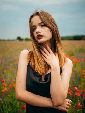 Bulutlu bir yaz gününde gelincik tarlasına poz veren siyah bir gece elbisesi içindeki güzel bir kızın portresi. Açık havada bir kadın modelin portresi. Yağmurlu bir hava. Gri bulutlar. Dikey çekim.