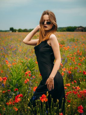 Siyah bir gece elbisesi ve güneş gözlüğü takmış bulutlu bir yaz gününde gelincik tarlasında poz veren güzel bir genç kız. Açık havada bir kadın modelin portresi. Yağmurlu bir hava. Gri bulutlar. Dikey çekim.