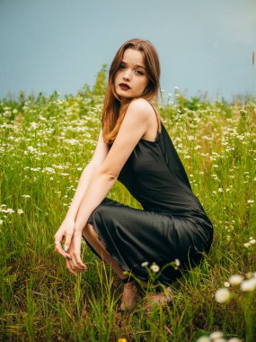 Siyah bir gece elbisesi içindeki güzel genç kız bulutlu bir yaz gününde papatya tarlasının önünde çömelmiş poz veriyor. Açık havada bir kadın modelin portresi. Yağmurlu bir hava. Gri bulutlar. Dikey çekim.