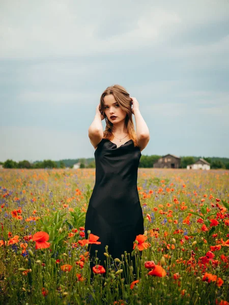 stock image Beautiful young girl in a black evening dress posing against a poppy field on a cloudy summer day. Portrait of a female model outdoors. Rainy weather. Gray clouds. Vertical shot.