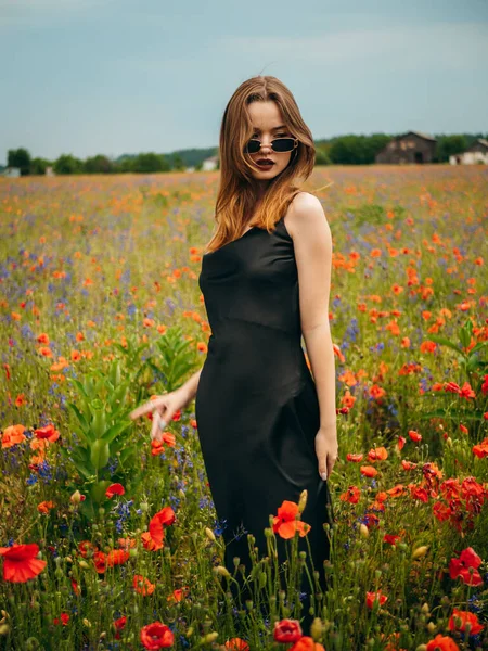 stock image Beautiful young girl in a black evening dress and sunglasses posing against a poppy field on a cloudy summer day. Portrait of a female model outdoors. Rainy weather. Gray clouds. Vertical shot.