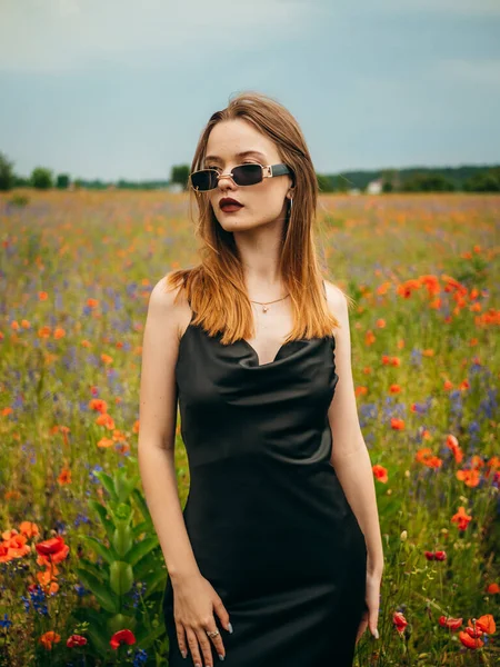 stock image Beautiful young girl in a black evening dress and sunglasses posing against a poppy field on a cloudy summer day. Portrait of a female model outdoors. Rainy weather. Gray clouds. Vertical shot.