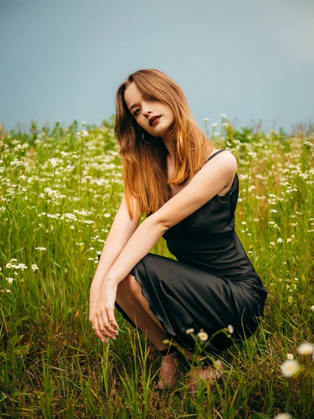 Beautiful young girl in a black evening dress poses crouching in front of a daisy field on a cloudy summer day. Portrait of a female model outdoors. Rainy weather. Gray clouds. Vertical shot.
