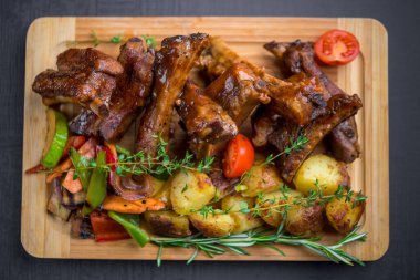 Kaburga manzarası. Patates, otlar ve ahşap tahtada servis edilen ızgara sebzelerle barbekü.