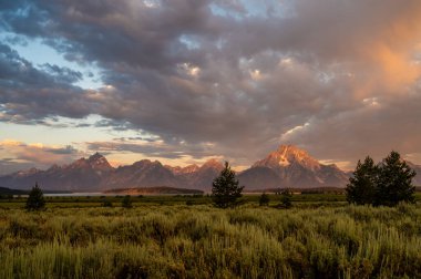 Gün doğumu Büyük Teton Dağlarının Bulutlarını ve Tepelerini aydınlatır adaçayı tarlasının arkasında