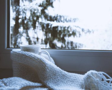 Bir kupa sıcak çay ve dışarıdaki kar manzarasına karşı cam kenarında yün örgü..