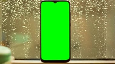 Cep telefonu, yeşil ekranlı damlacıkları olan bir pencere camına dayanıyor.